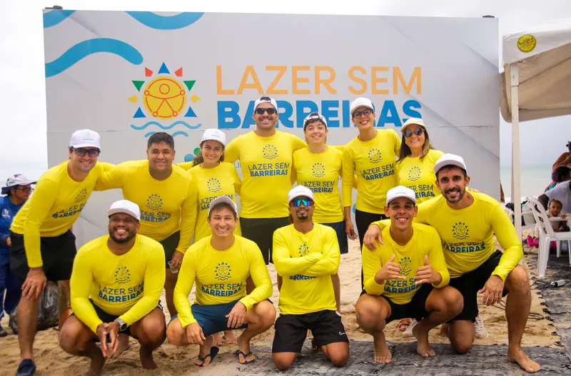 Equipe de voluntários de camisa amarela do Lazer sem Barreiras reunida na praia, em frente ao painel do projeto