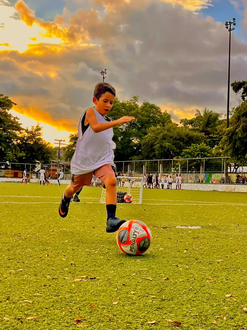 Menino correndo para chutar a bola durante treino de futebol no Gramadão da Campina do Barreto, ao pôr do sol
