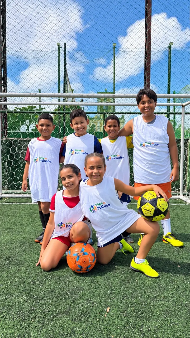 Seis crianças de colete do Time da Inclusão posando com bolas em frente ao gol, no treino de futebol