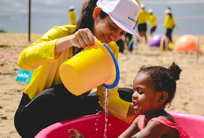 Voluntária brincando com água e uma menina numa piscininha, durante o Lazer sem Barreiras na praia